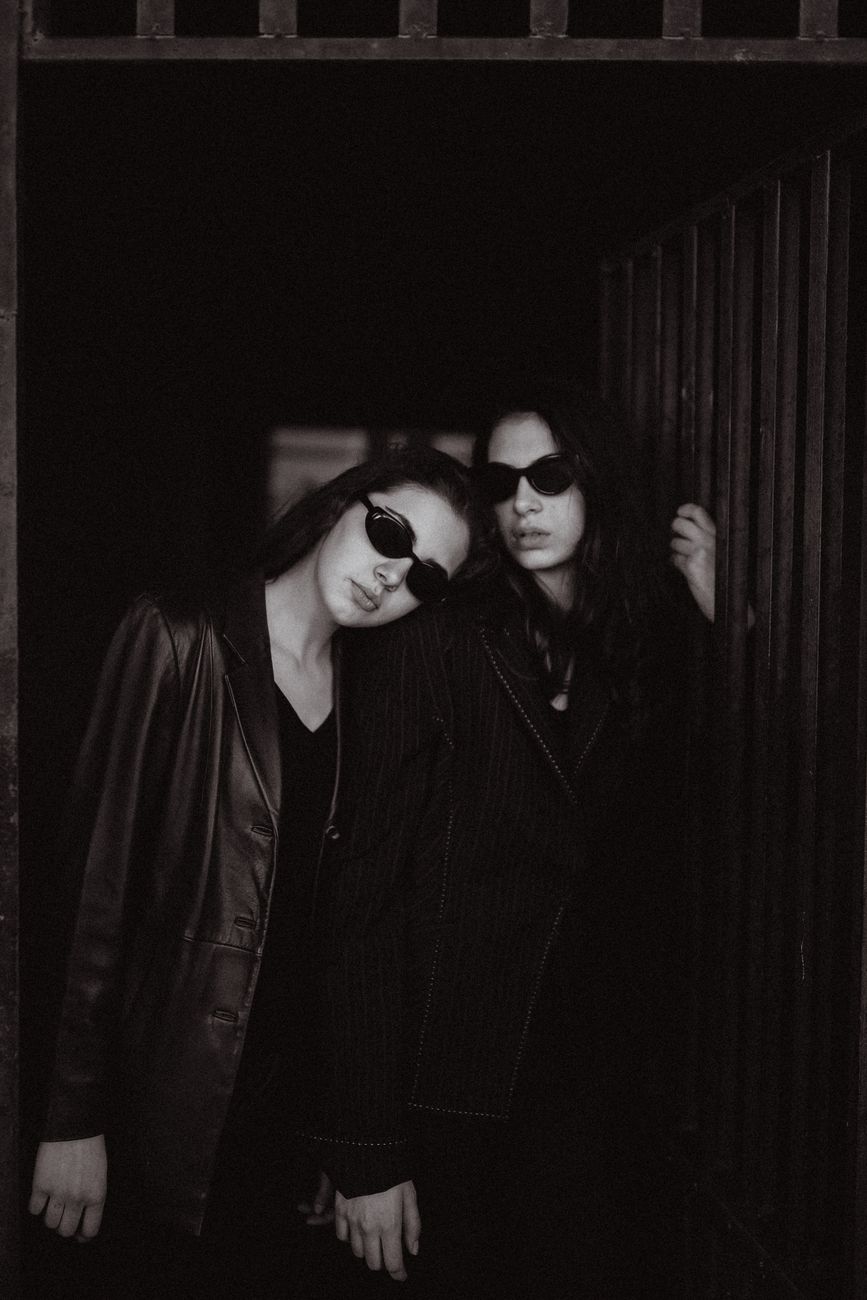 Two models in dark sunglasses leaning together in a doorway